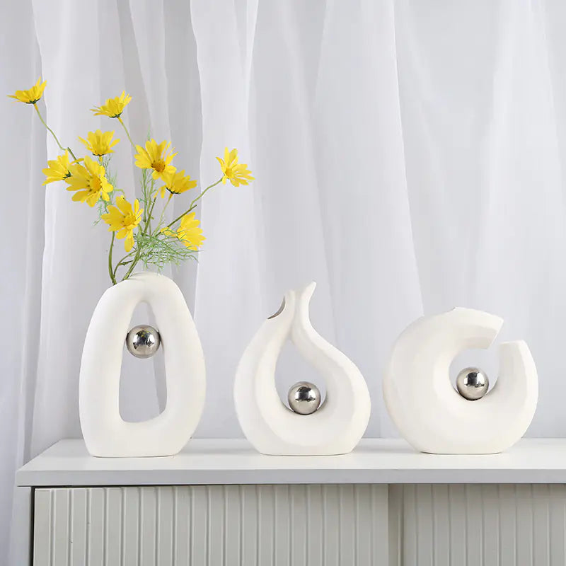 Three white decorative vases with silver spheres on a white surface against a white curtain background