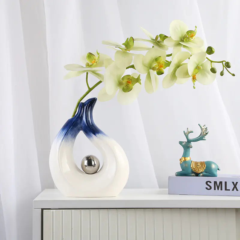 Decorative vase with a blue and white gradient, silver sphere, and white orchids on a white surface.