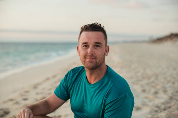 Man in teal shirt sitting on a beach with ocean and sky in the background