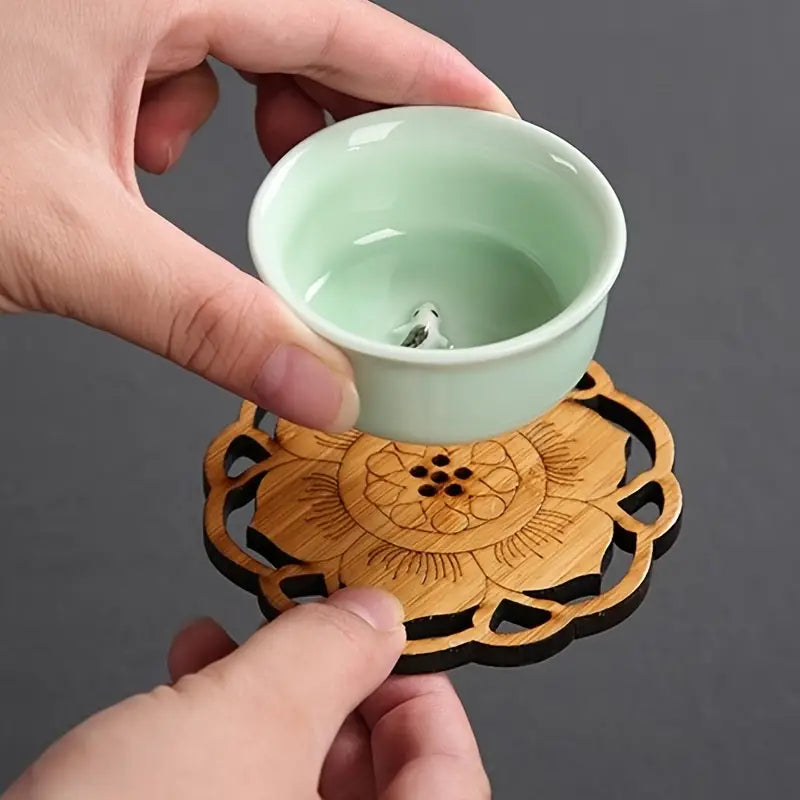 Hand holding a light green ceramic cup on a wooden coaster with floral design against a dark background